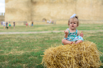 Naklejka na ściany i meble A little girl sits on a straw in a dress and a bow near the wall of the fortress