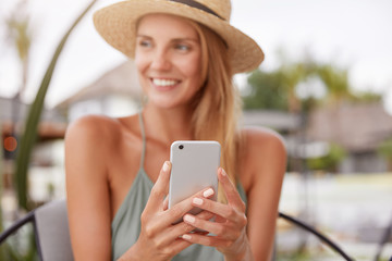 Cheerful relaxed woman uses smart phone for chat with friends, sits in modern coffee shop or terrace cafe. Pretty female reads good news on internet website, enjoys summer rest. Focus on cell phone