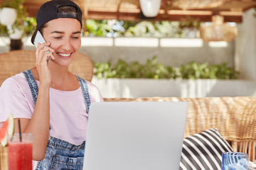 Horizontal shot of happy young female in stylish cap looks joyfully into screen of laptop, makes online project, connected to wireless internet, has conversation with someone on smart phone.