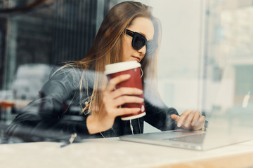 Young girl sit in coffee place in front of the window look at her laptop and drink tea from red cup