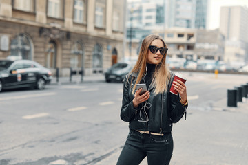 Pretty girl stand on the street listen music from her headphones and drink coffee, windy weather