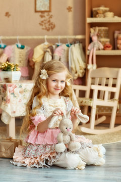 Cute smiling girl with light long hair in a vintage dress with a plush Bunny sits on the floor in the nursery.