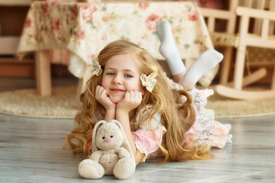 Cute smiling girl with light long hair in a vintage dress with a plush Bunny lies on the floor in the nursery.