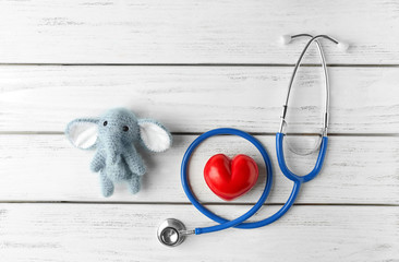 Cute knitted elephant, small heart and stethoscope on white wooden background. Concept of visiting children's doctor