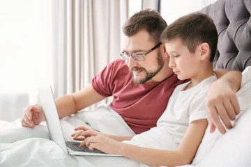 Little boy and his dad using laptop in bedroom