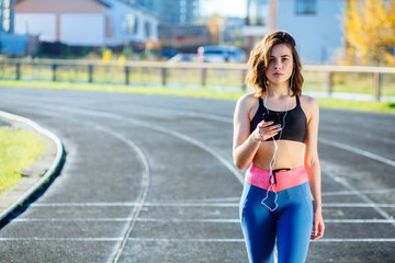 Purposeful european runner female listening to running motivation music with earbuds on her mobile phone app for run on track in athletics stadium. Athlete enjoying fitness active life concept.