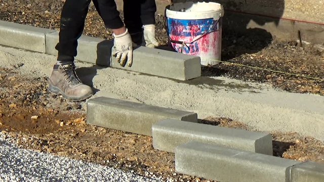 worker installing concrete curb stone and using string with metal stakes to level at sidewalk construction site.