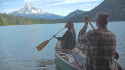 Young loving couple rowing away on a boat together with some paddles on a lake. she looks back at him, and smiles.