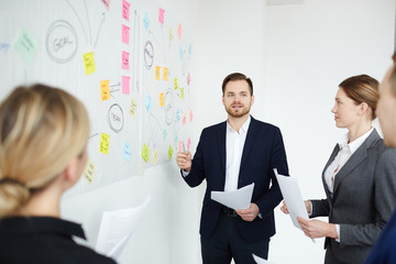 Confident businessman making report or presentation by whiteboard during start-up meeting with colleagues