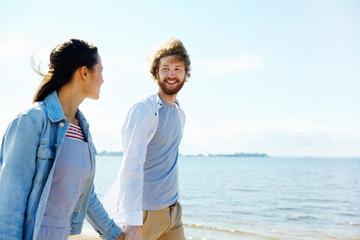 Happy young man looking at his wife and holding her by hand while both walking by waterside