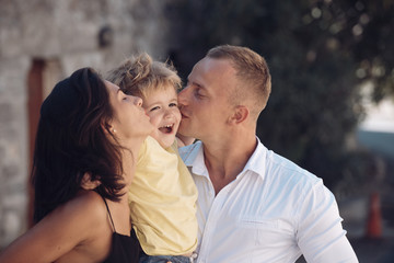 Happy family spend time together, urban background. Parents kissing son in cheeks, happy boy with smiling face. Mother and father love their child. Happy family concept. Parenthood, hugs, love, kiss.