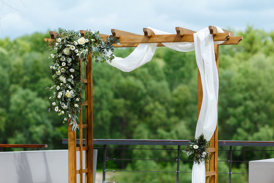 Area of the wedding ceremony near river on the pier. Wooden rectangular arch, white chairs decorated with flowers, greenery, petals, eucalyptus and tulle. Cute, trendy rustic decor