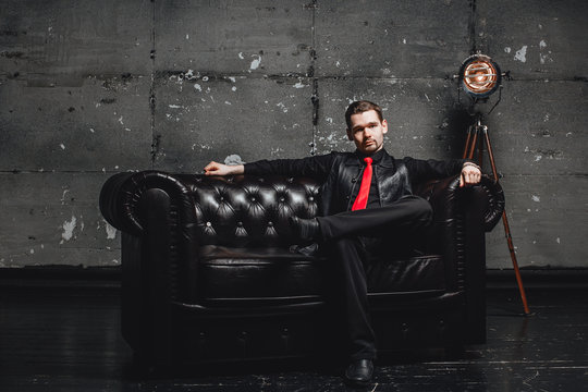 Handsome businessman in black suit sitting on sofa and looking away. Young man looking like executive or head of company or enterprise. Room in loft style.