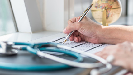 Doctor writing on medical health care record, patients discharge, or prescription form paperwork in hospital clinic with physician's stethoscope on office desk