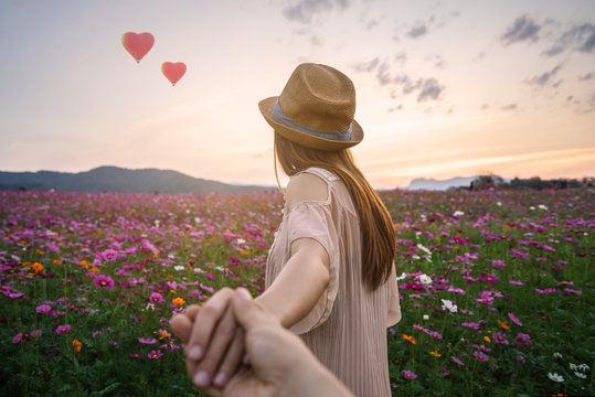 Young woman traveler holding man's hand and looking heart shape balloons flying in the sky on flowers field, Couple vacation travel concept