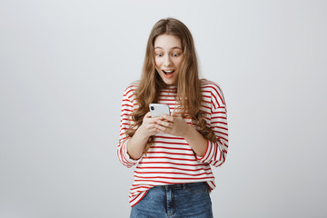 Dreams come true with technology help. Portrait of trendy modern woman with long blond hair holding smartphone, looking with amazement at screen, happy to receive reply from classmate over gray wall