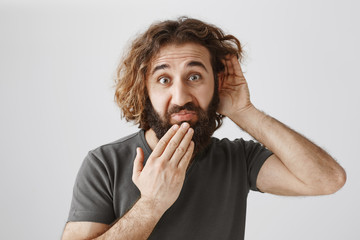 What a shameful story. Portrait of displeased surprised man with curly hair holding hands near ear and mouth, eavesdropping conversation, hearing unpleasant gossip, standing over gray background