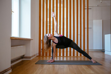 European woman practicing yoga, standing in Revolved Triangle exercise, Parivrrta Trikonasana pose, working out wearing sportswear, indoor full length, against wooden studio background