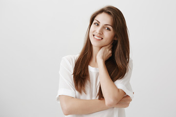 Girl feels discomfort talking with strangers. Indoor portrait of charming shy caucasian student touching neck and smiling politely, feeling annoyed but hearing out boring stories over gray wall
