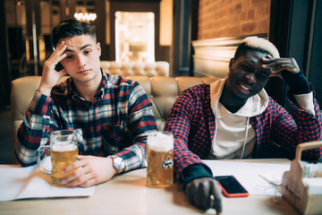 Handsome men's friends are sitting upset while drinking beer and cheering for sport team at the pub