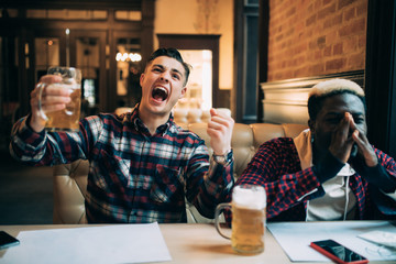 Handsome young man holding his beer and happy shouting looking at the TV screen watching football game with his upset afro american friends at the local pub while support different teams.