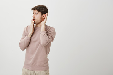 Astonished and surprised handsome male model turned left while holding hands near ear and opened...