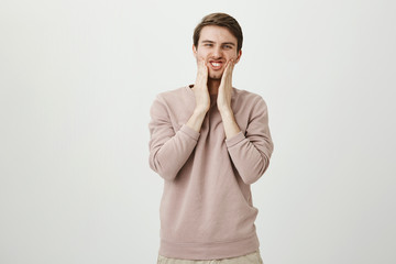 Portrait of funny handsome caucasian male squeezing cheeks with both hands and squinting at camera, standing against gray background. Guy looks in mirror and thinking that it is time to shave