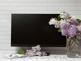 Beautiful white and violet lilac flowers bouquet in glass vase with opened note-book and black monitor screen on the white brick and wooden background, mock up front view