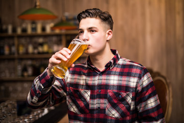 Young attractive man in casual clothes is drinking beer while sitting at bar counter in pub