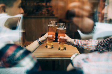 Close up hands of friends sitting at the pub cheers with beers glasses trough window.