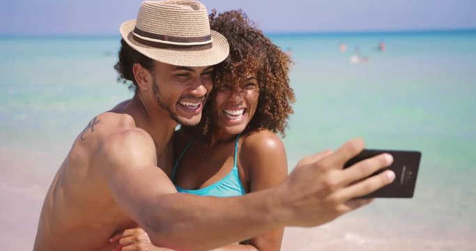 Cheerful young happy ethnic man and woman standing on sunny sandy beach and taking selfie together.