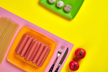 Sausages with tomatoes, spaghetti and garlic on a yellow and pink minimal background. Flat lay. Top view.