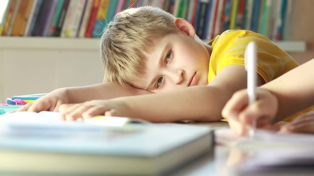 Elementary schoolboy looking tired and exhausted resting his head on desk in classroom.