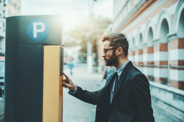Bearded serious man entrepreneur in eyeglasses and a formal suit is using parking pay station terminal; handsome businessman in glasses and with the beard paying his parking time via automatic kiosk
