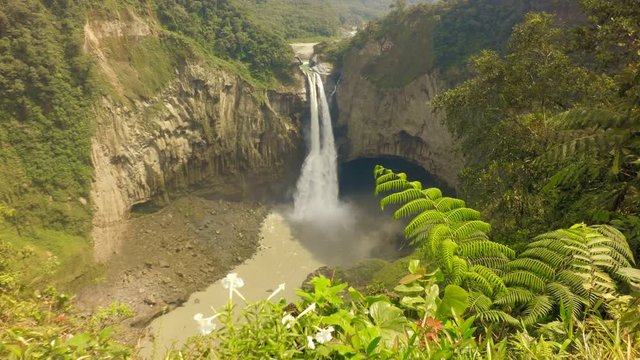 A wide shot of the San Rafael waterfall plunging into a rocky canyon in Ecuador, framed by dense tropical vegetation and ferns, a remote verdant mountain landscape under bright daylight.