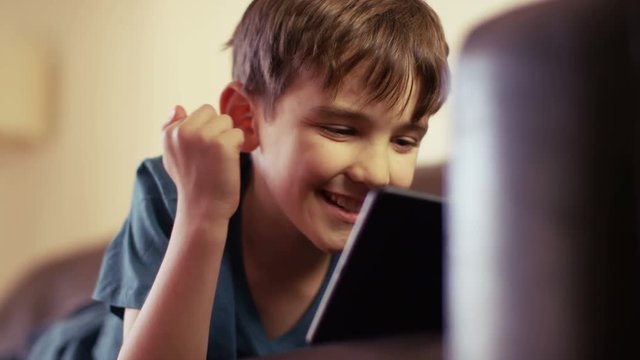 A very happy young boy playing on his tablet while lying down on couch at home.