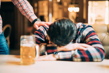 Young man in casual clothes is sleeping near the mug of beer on a table in pub, another man is...