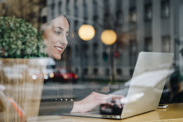 beautiful brunette woman sitting alone in cafe near the window and working on laptop. attractive fashion blogger writing a new blog post on her computer while drinking coffee. shot through the window