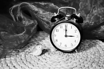 Black and white picture of old alarm clock showing 3 o clock with two bells standing on table with pattern place mat and a little china chicken (porcelain), change of time, early bird