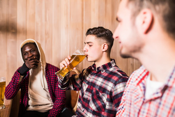 Three multiracial men in casual clothes are smiling and drinking beer while sitting at bar counter in pub