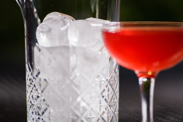 A small low glass with a red alcoholic cocktail is on the table next to a glass vase full of large square pieces of clear ice, against a background of beautifully blurred green plants.