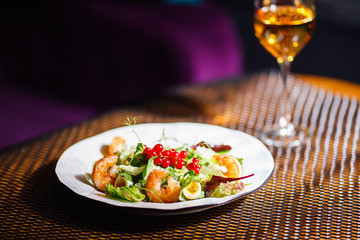 A plate with a beautiful appetizing salad of fresh vegetables, shrimps, quail eggs and lingonberries is on the table with a drink in a wine glass.