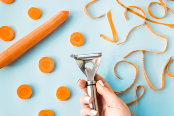 Carrot cleaning with a special kitchen appliance in female hand. On a blue background. Bright carrot shavings and rings. Top view, flat lay