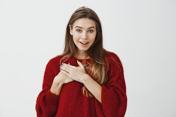 Studio portrait of pleased flattered young attractive woman in red loose sweater, holding palms on chest and smiling broadly, speaking with boyfriend who confessing in love over gray background