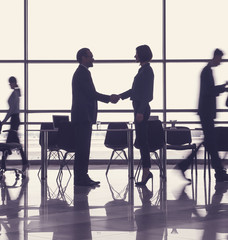 Man and woman standing in workspace and shaking hands. People walking on background