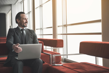 Jolly solid man sitting on lounge with notebook, he is looking at window and laughing. Copy space...