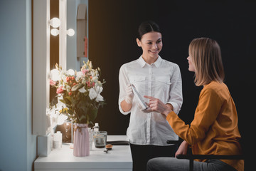 Outgoing visagiste presenting cosmetic product for cheerful client. Discussion during job concept