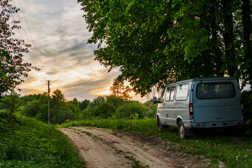 The minibus stands on the side of a dirt road. The machine stands under large linden trees. From behind you can see the sky and the sunset.