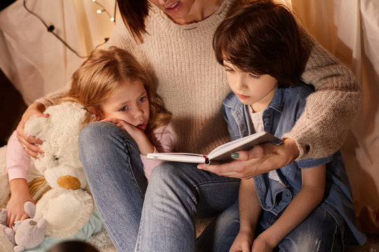 Mother sitting and hugs kids. They in comfortable nursery reading interesting story for night. Concept of childhood