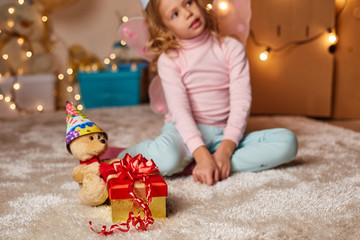 Joyous girl sitting on carpet in gaming room with teddy bear and beautiful present box